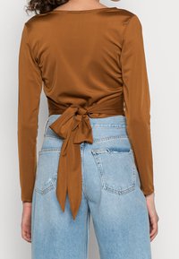 Top lungo a maniche con tessuto in satin marrone, caratterizzato da un design con nodo sulla schiena. Abbinato a jeans in denim chiaro, usurati.
