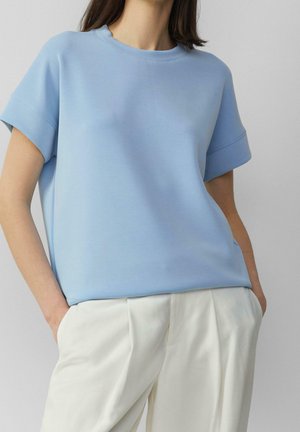 Kobieta ubrana w jasnoniebieską bluzkę z krótkim rękawem, wpuszczoną w wysokowalne, białe plisowane spodnie, z rękami w kieszeniach.