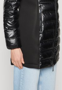 Sort polstret jakke med skinnende materiale på arme og krop, med mat sort stof på siderne og lommerne. Lynlås lukning.