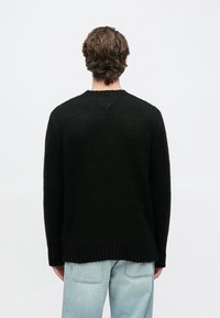 Μαύρο πλεκτό πουλόβερ με υφημένη υφή, στρογγυλό λαιμόκοψη, ριμπ μανσέτες και τελείωμα. Φορεμένο πάνω από ανοιχτόχρωμο τζιν.