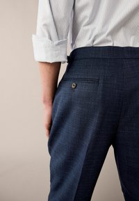Marineblaue Hose mit strukturiertem Muster, ausgestattet mit einer einzelnen Gesäßtasche, die mit einem braunen Knopf gesichert ist. Sichtbarer, weiß gestreifter Hemdärmel mit Bündchen.