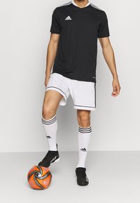Mužský fotbalista v černém dresu Adidas, bílých šortkách a ponožkách ovládá oranžový a černý fotbalový míč nohou.