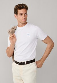 T-shirt blanc à manches courtes avec un logo bleu marine subtil sur la poitrine, associé à un pantalon crème et un pull en tricot beige drapé sur l'épaule.