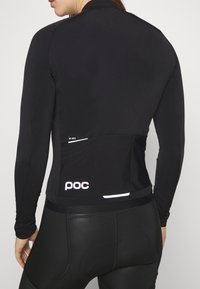 Femme portant un maillot de cyclisme noir à manches longues et un cuissard rembourré, vue de dos, avec le logo "poc" sur la poche du maillot.