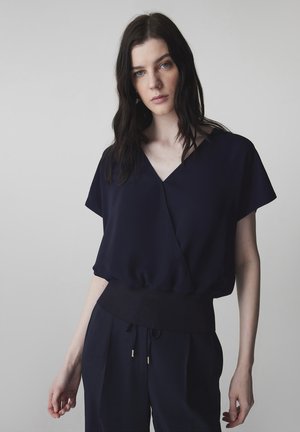 Mujer con cabello largo y oscuro que lleva una blusa negra holgada con escote en V, cinturilla ancha y pantalones negros a juego, sobre un fondo liso.