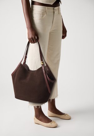 Sac fourre-tout en suède marron avec des sangles en cuir et un accent doré. Porté avec un pantalon ample couleur crème et des ballerines beiges.