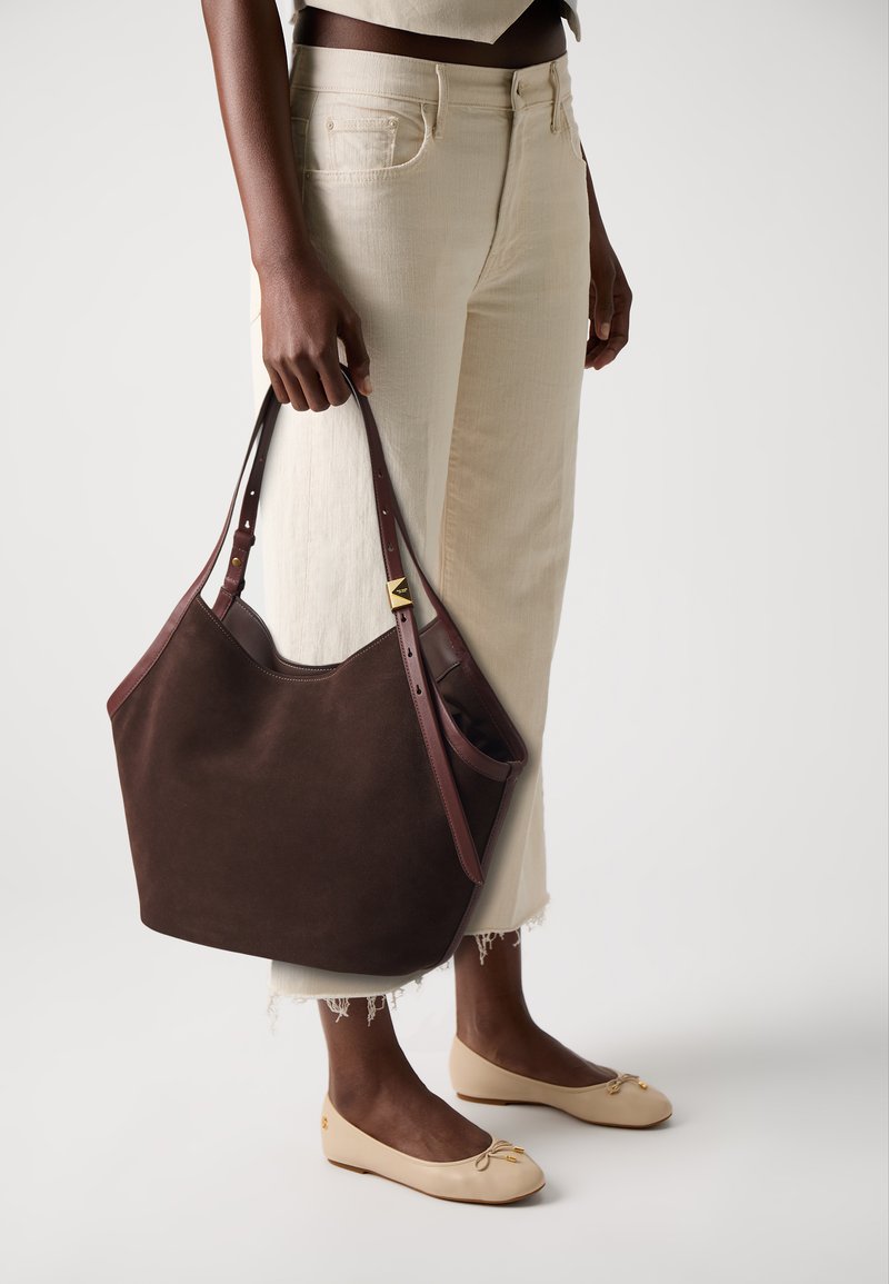 Borsa a spalla in suede marrone con manici in pelle e un accento dorato. Indossata con pantaloni a gamba larga color crema e ballerine beige.