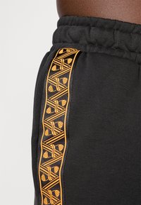 Un pantalon de survêtement noir avec une taille élastique présente une bande latérale aux motifs géométriques orange complexes. Le tissu semble lisse et doux.
