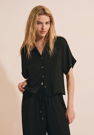 Mujer con cabello rubio largo que lleva una camisa negra de manga corta con botones y pantalones negros con cordón en un fondo liso.