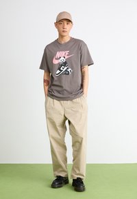 Grå t-skjorte med en rosa Nike-logo og panda-grafikk, sammen med lyse beige bukser og svarte sko. En enkel caps fullfører antrekket.