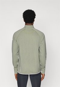 Világoszöld, texturált, hosszú ujjú dzseki magas gallérral, gyémántmintával, oldalsó hasítékokkal és kontrasztos fehér részletekkel.