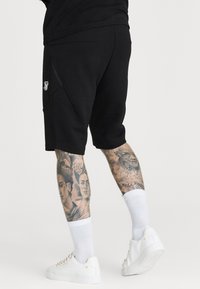 Musta urheilu(shortsit), joissa on sivutaskut ja pieni valkoinen logo. Yhdistetty valkoisiin lenkkareihin ja polviin yltäviin valkoisiin sukkiin.