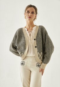Cárdigan de punto en patrón de zigzag en beige y negro, con un corte holgado, mangas amplias y dos botones. Combinado con pantalones de color claro con bordados florales.