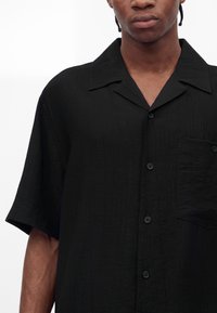 Jeune personne à la peau foncée portant une chemise noire à manches courtes avec col et poche poitrine, montrée de la moitié de la poitrine jusqu'en haut.