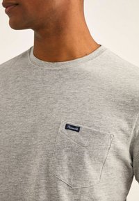 Grijze katoenen T-shirt met een ronde hals en een patch pocket met een marineblauw label. Zachte textuur, korte mouwen en een minimalistisch ontwerp.