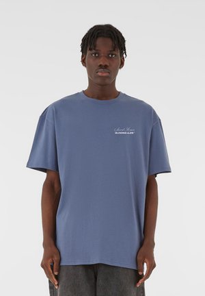 Tricou oversized din bumbac albastru, cu guler rotund și mâneci scurte, având un logo alb pe piept și o textură netedă, moale.