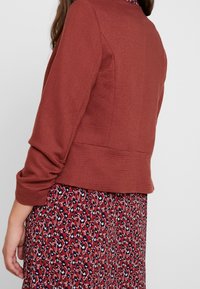 Rust-colored ribbed jacket with three-quarter sleeves, paired with a red dress featuring a floral pattern in navy and white.