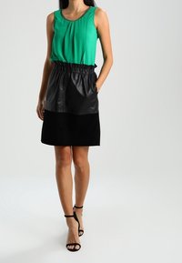 Top verde senza maniche con pieghe, abbinato a una gonna in pelle nera e tessuto. L'outfit presenta una vita arricciata e décolleté con cinturini alla caviglia.