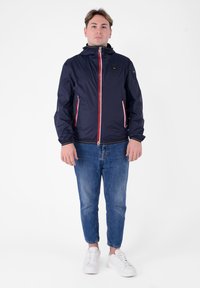 Giacca a vento navy con dettagli rossi sulle zip e tasche laterali, dotata di cappuccio e polsini elasticizzati, abbinata a jeans blu e sneakers bianche.
