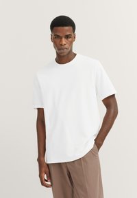 Weißes T-Shirt mit kurzen Ärmeln, runder Ausschnitt, glatte Textur, mit subtiler Markenkennzeichnung auf der Vorderseite. Kombiniert mit hellbraunen Hosen.