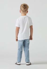 T-shirt blanc à manches courtes, jean en denim bleu clair et chaussures rayées noir et blanc. Vue de dos d'un enfant avec une coiffure courte et épi.