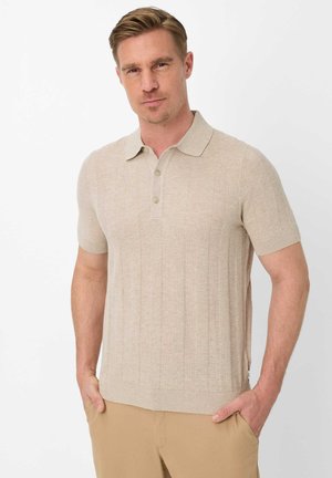 Mann mit kurzen blonden Haaren, der ein beigefarbenes, kurzärmeliges Strick-Poloshirt und beige Hosen trägt, steht mit den Händen in den Taschen.