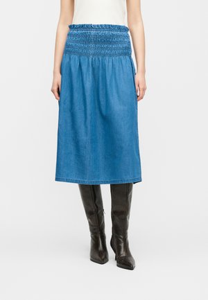 Femme portant une jupe midi bleue avec une taille froncée et des bottes noires à talons hautes jusqu'aux genoux, debout devant un fond uni.