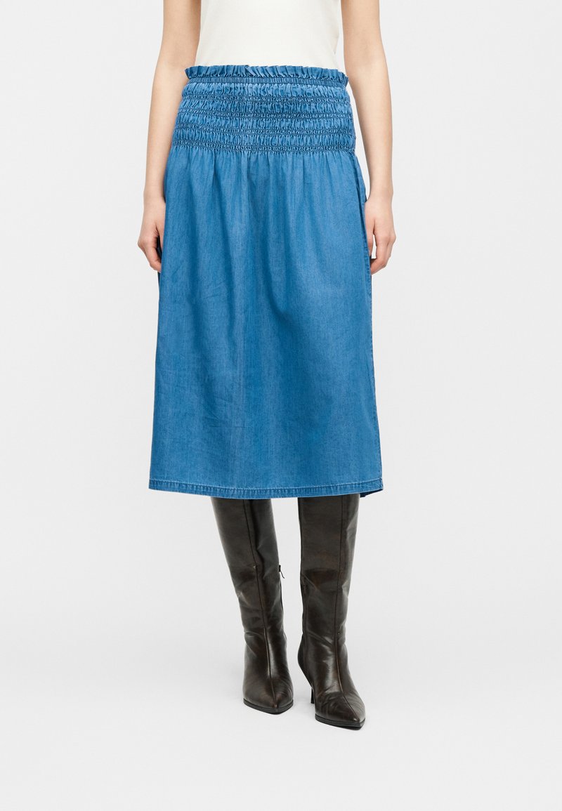 Γυναίκα με μπλε μίντι φούστα με πιέτες στη μέση και μαύρες μπότες με τακούνι μέχρι το γόνατο, στέκεται μπροστά σε απλό φόντο.