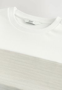 T-shirt blanc avec une rayure horizontale beige texturée ; présente un col rond et un tissu doux. L'étiquette indique "NEXT, TAILLE STANDARD."