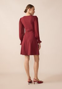 Robe rouge avec corsage ajusté, manches à volants transparentes et jupe évasée. Associée à des escarpins rouges à lanières arrière avec un détail en nœud.