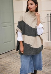 Beige and gray knit poncho with a high collar, featuring a wavy pattern; paired with a white shirt and denim skirt. Smooth texture.