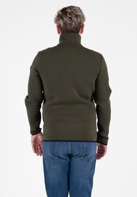Pullover verde scuro a maniche lunghe con colletto alto, texture liscia e design aderente; abbinato a jeans in denim blu.