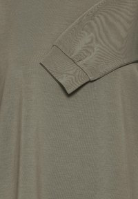 Ilgų rankovių drabužis nykstančios alyvuogių žalios spalvos, su matomais rankogaliais ir minkšta, tekstūruota medžiaga.