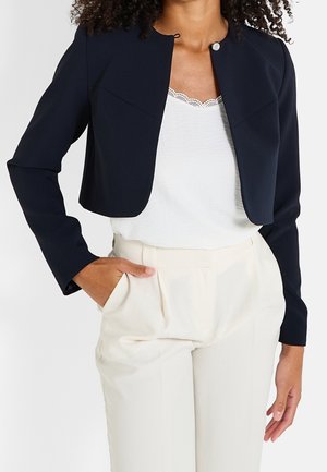 Femme portant une veste noire courte par-dessus un haut blanc bordé de dentelle et un pantalon crème taille haute, main dans la poche.