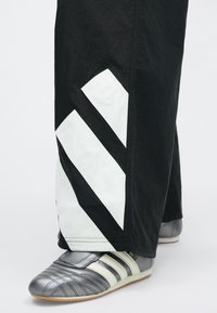 Schwarze Hose mit weißen diagonalen Streifen, kombiniert mit grauen Sportschuhen mit weißen Streifen und einem Klettverschluss, die ein elegantes Design zeigen.