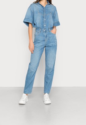 Kobieta nosząca jasnoniebieską koszulę dżinsową i dżinsy z wysokim stanem, w białych trampkach, stojąca z jedną ręką w kieszeni na tle jednolitego tła.