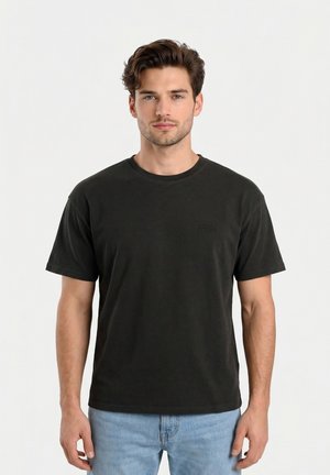 Giovane uomo con capelli corti e mossi, che indossa una maglietta nera semplice e jeans azzurri, in piedi davanti a uno sfondo bianco.