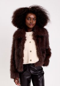 Veste en fausse fourrure brune avec un col châle, associée à un pull crème côtelé avec des boutons décoratifs, et un pantalon en cuir noir.