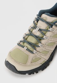 Light tan hiking shoe with olive green mesh panels, blue and white laces, and a black rubber sole. Features textured surface and cushioned collar.