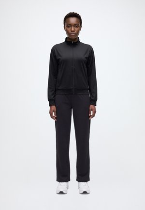 Un mannequin se tient devant un fond uni, vêtu d’une veste noire zippée et d’un pantalon assorti, avec des baskets blanches.
