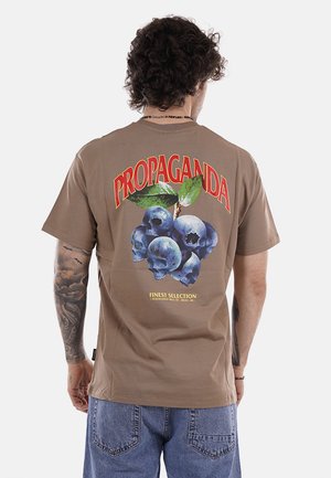 Uomo con capelli ricci e tatuaggi che indossa una maglietta marrone con mirtilli fatti di teschi e la scritta "PROPAGANDA" sul retro.