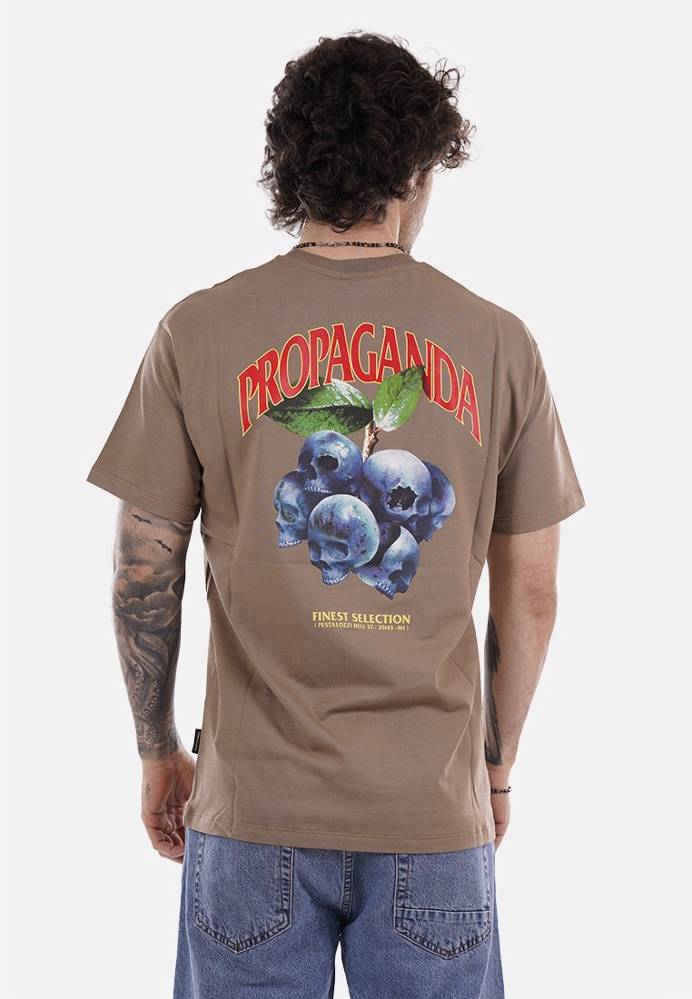 Uomo con capelli ricci e tatuaggi che indossa una maglietta marrone con mirtilli fatti di teschi e la scritta "PROPAGANDA" sul retro.