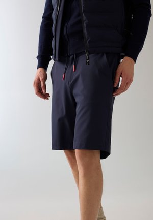 Homme portant un short bleu marine avec des embouts rouges aux cordons et une veste bleu marine, debout devant un fond clair uni.