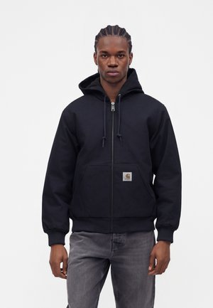 Ung mand med fletninger iført en sort hættetrøje med lynlås og grå jeans, stående foran en hvid baggrund.
