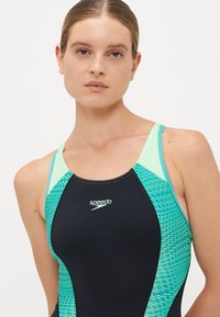 Mlada ženska s svetlo kožo in zvezanimi lasmi, oblečena v črno in smaragdno modro kopalke Speedo, na enobarvni svetli ozadju.