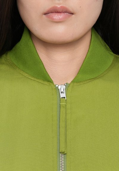 Green zip-up jacket with a high collar, featuring a silver zipper and smooth fabric texture. Close-up of the neckline and zipper area.