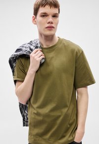 Olivno zelena bombažna majica s kratkimi rokavi. Na prsih subtilen logotip. Črno-bela karo flanelasta srajca je prevrnjena čez eno ramo.