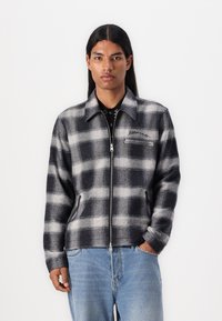 Black and white plaid jacket with a zip closure, long sleeves, and chest pocket; soft, textured fabric. Paired with light blue jeans.