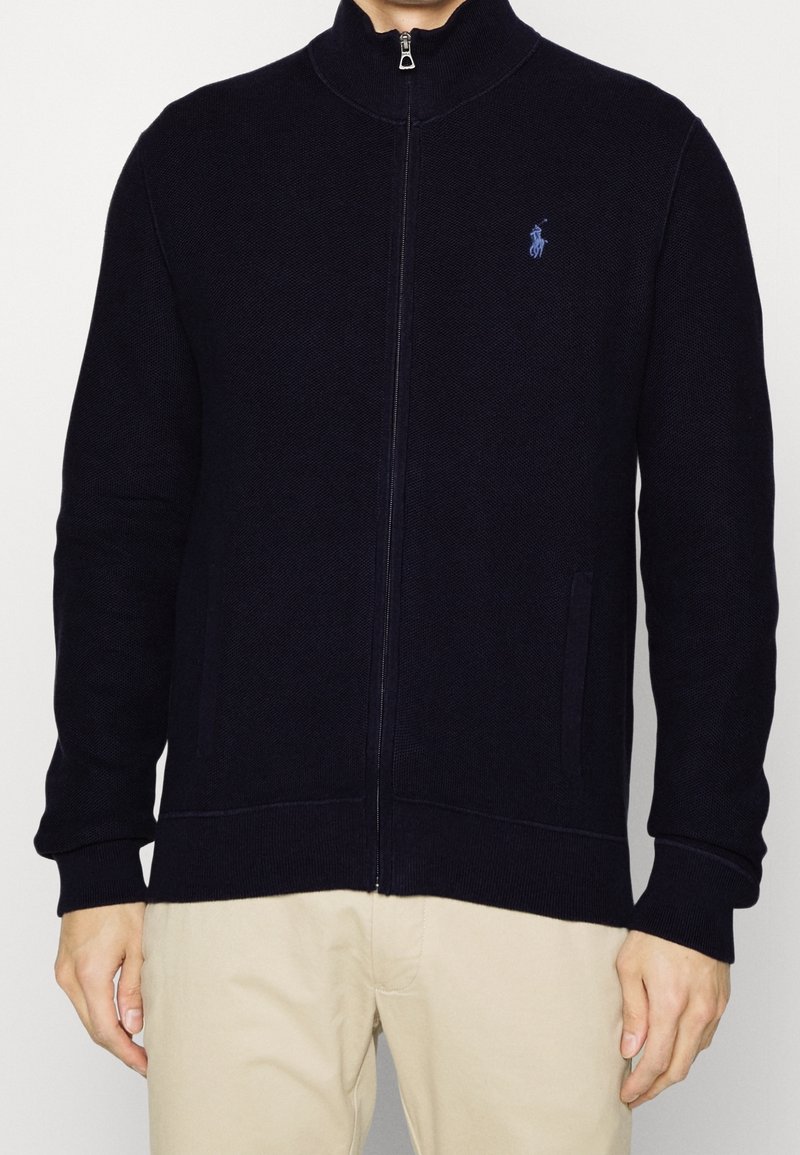 Laivastonsininen vetoketjulla varustettu takki, jossa on korkea kaulus, resorit hihansuissa ja helmassa. Vasemmassa rinnassa pieni kirjailtu logo ja sivutaskut.