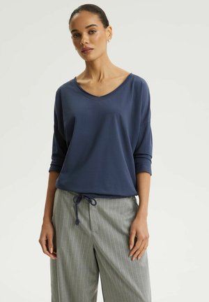 Kobieta ubrana w granatowy top z dekoltem w serek i rękawami do łokcia oraz szare spodnie w paski, zawiązane w talii, stojąca na jednolitym tle.